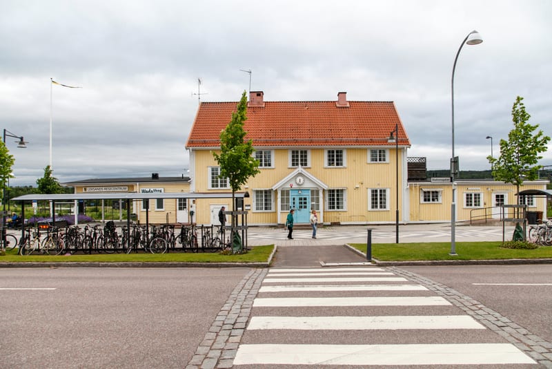 En gul timmerbyggnad med rött tak står centralt i bilden. Byggnaden har många fönster med vita karmar. Framför byggnaden finns en blå entrédörr med en skylt ovanför som visar klockan. Ett ungt träd står framför dörren. Framför byggnaden, till höger,