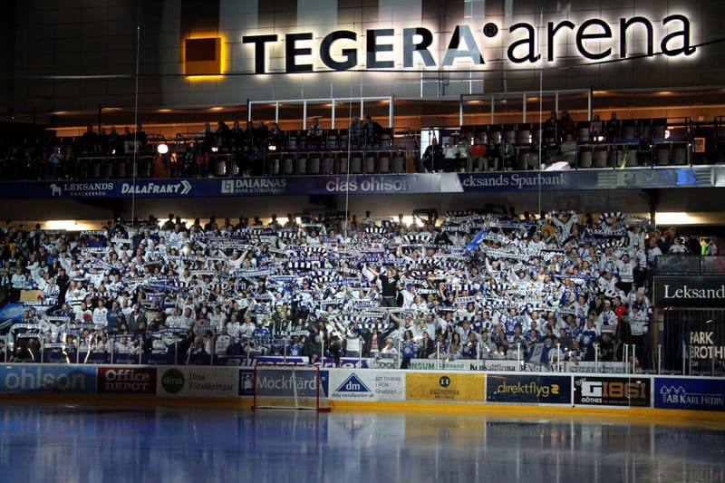 Bilden visar interiören av en ishockeyarena. I förgrunden syns isen och en del av sargen med reklamskyltar. På sargen finns sponsorlogotyper som "ohlson", "Office DEPOT", "Dina Försäkringar" och "Mockfjärd". En målbåge syns också nära isen. Bakom sar