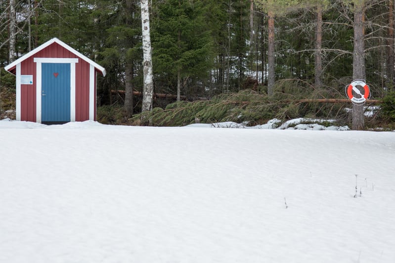Bilden visar ett rött uthus med en blå dörr och vita knutar, omgivet av snö. På dörren syns en hjärtformad utskärning. Till vänster om dörren sitter en liten vit skylt. Framför uthuset breder ett snötäckt fält ut sig. I bakgrunden finns en skog med b