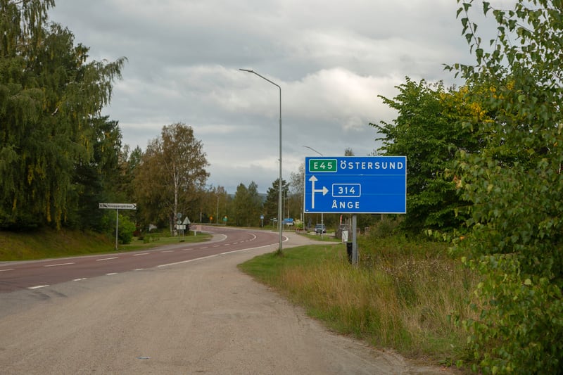 Denna bild visar en vägkorsning i ett lanskap med träd och gräs. I förgrunden syns en grusväg som möter en asfalterad väg. Den asfalterade vägen slingrar sig vänsterut och har röda körfält med vita heldragna linjer. Längs vägen finns en blå vägskylt