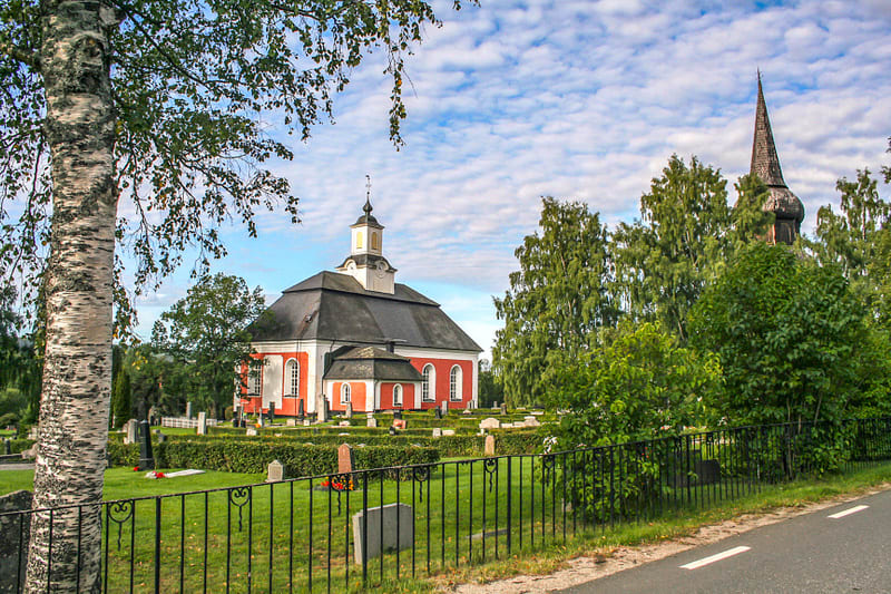 Bilden visar ett kyrkobyggnad med röda väggar och vita fönsterkarmar, med ett svart tak och en vit tornspira. Till höger om kyrkan syns en annan spira, delvis skymd av lövträd. Framför kyrkan ligger en kyrkogård med gräsmattor, gravstenar och en häck