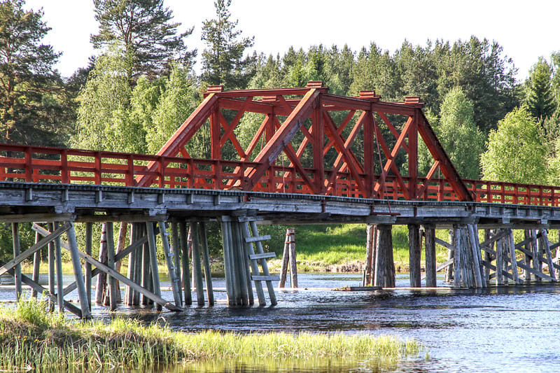 Bilden visar en korsning av en röd fackverksbro och en träbro över ett vattendrag. Den röda fackverksbron utgör den övre delen, med ett rött räcke som avgränsar gångbanan. Nedanför den röda bron syns en robust träbro som bärs upp av vertikala och dia