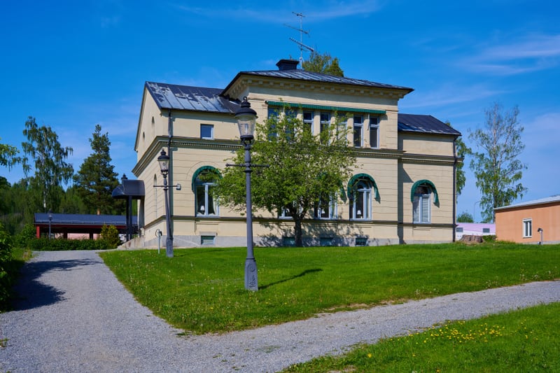 Bilden visar en gulbeige byggnad med mörkt tak, belägen i ett grönområde under en klarblå himmel. Byggnaden har flera fönster, där några har bågformade överliggare i en grönblå nyans, och andra är rektangulära. En liten topptanna och antenner syns på