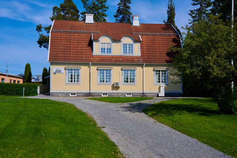 Bilden visar en gul trähusbyggnad med brun-orange tegeltak under en klarblå himmel. Huset har vita fönster med spröjs och fönsterluckor. På framsidan av byggnaden finns en skylt med texten "PASTORS EXPEDITION" och en vit utomhusenhet som kan vara en