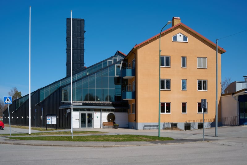 Bilden visar byggnader under en klarblå himmel. Till vänster finns en svart byggnad med glaspartier som lutar uppåt och en del av taket i vinkel. Byggnadens entré har vita dörrar och en skylt med texten "sportforumgatan". Framför entrén finns en bänk