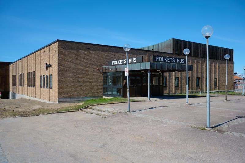 Bilden visar fasaden på en tegelbyggnad med texten "FOLKETS HUS" ovanför två ingångar. Ingångarna har glasdörrar och ett utskjutande tak med inbyggda lampor. Byggnaden är huvudsakligen gjord av gulbrunt tegel och har flera fönster längs sidorna. Take