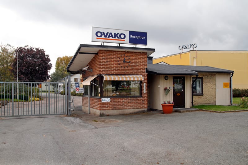 Bilden visar entréområdet till en anläggning, troligen en industri eller fabrik, med en byggnad som fungerar som reception. Byggnaden består av två delar: en mindre del i rött tegel med ett fönster och en randig markis, samt en större ljus byggnad me
