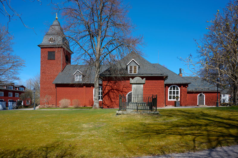 En röd träkyrka med ett mörkt tak och en klockstapel är centralt placerad i bilden under en klarblå himmel. Kyrkan har vita fönsterkarmar och takkupor. Framför kyrkan syns en gräsmatta som är delvis i skugga från träden. Ett grått stenmonument med et