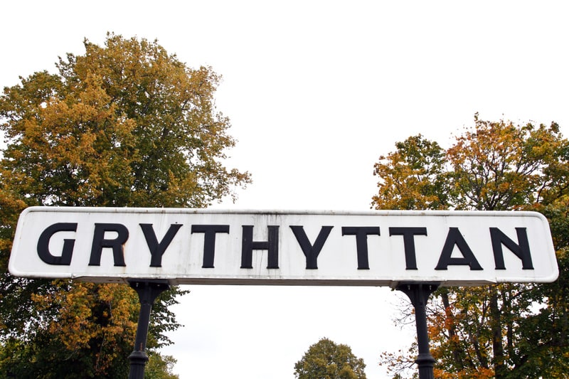 En vit skylt med svarta versala bokstäver som bildar ordet "GRYTHYTTAN" är centrerad i bilden. Skylten hänger mellan två svarta stolpar. Bakom och runt skylten syns träd med lövverk i skiftande gröna och gula/orangea höstfärger, vilket indikerar att