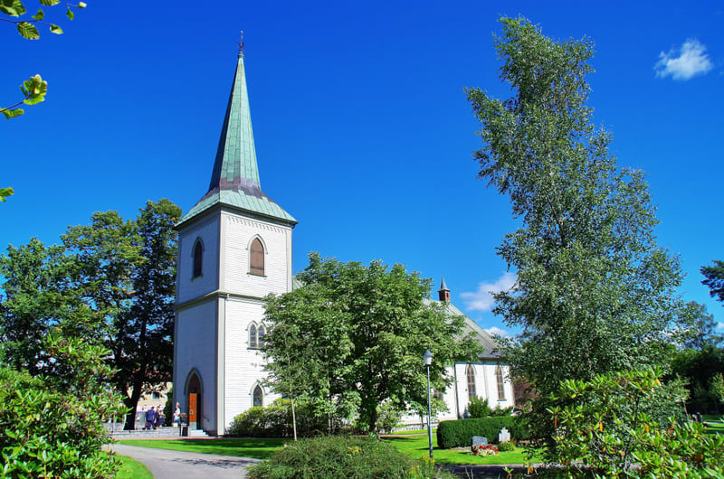 Bilden visar en vit kyrkobyggnad med ett högt, spetsigt grönt tak tornande upp mot nordväst. Fasaden är ljus och har flera fönster. Intill kyrkan finns en grön gräsmatta med flera träd och buskar som har gröna lövverk. I nederkanten syns en asfaltera