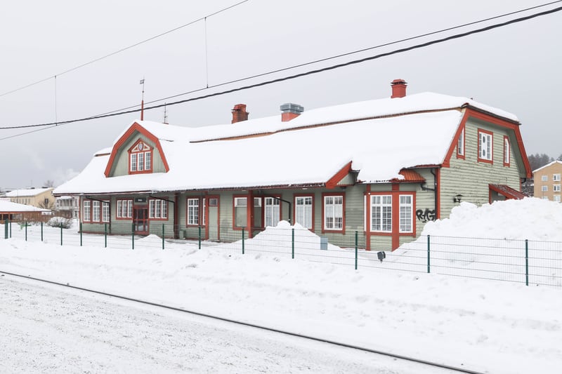 Bilden visar en medelstor ljusgrön träbyggnad med rött tak och vita fönsterkarmar, täckt av snö på taket och runt omkring. Byggnaden har flera skorstenar, en takkupa och en entré med en dörr och fönster. En grå himmel syns i bakgrunden. Framför byggn