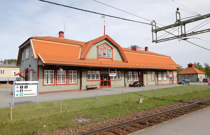 En bild visar en tågstation med ett stort trähus med orange takpannor och gröna väggar med vita fönsterkarmar. Huset har flera fönster och dörrar samt en centralt placerad takkupa med fönster. Framför byggnaden finns en asfalterad plattform med bänka