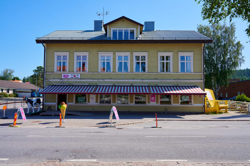 En gulmålad träbyggnad med vita fönsterkarmar och ett mörkgrått plåttak syns i bildens centrum. På markplan finns en markis med röda och vita ränder som täcker skyltfönstren. Utanför byggnaden, till vänster från betraktarens perspektiv, står en svart