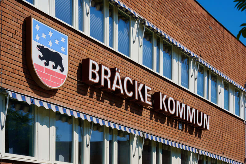 En bild visar fasaden på en tegelbyggnad med texten "BRÄCKE KOMMUN" i stora vita bokstäver monterade på väggen. Till vänster om texten sitter en skylt med Bräcke kommuns vapen, föreställande en svart björn på en bakgrund med blått fält med vita stjär