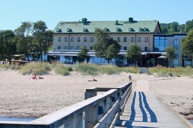Bilden visar en strandmiljö med en träbrygga i förgrunden som sträcker sig ut mot vattnet. Bryggan är av trä och har en avgränsning med brädreläer på sidorna. I bakgrunden syns en bred sandstrand där det bland annat ligger en person på en handduk. Ba