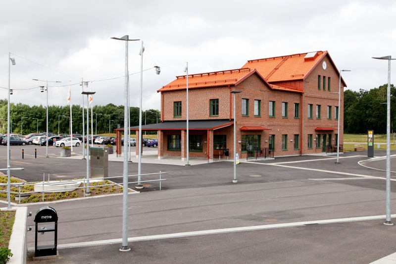 Bilden visar en tegelbyggnad med rött tegeltak och mörkgröna fönsterramar. Byggnaden har två våningar och en utskjutande del med tak och pelare på markplan. Framför byggnaden finns ett asfalterat område med parkeringsplatser och flera metallstolpar m