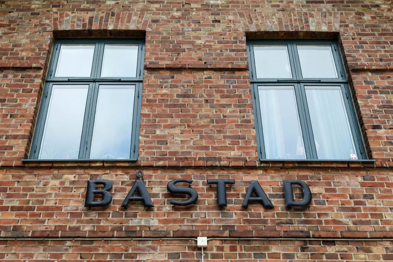 Fasaden på en byggnad sedd snett underifrån visar två fönster med mörkgrå ramar, placerade i en röd tegelvägg. Över varje fönster finns en välvd tegelkonstruktion. Under fönstren, längs tegelväggen, sitter stora, svarta bokstäver som formar ordet "BÅ