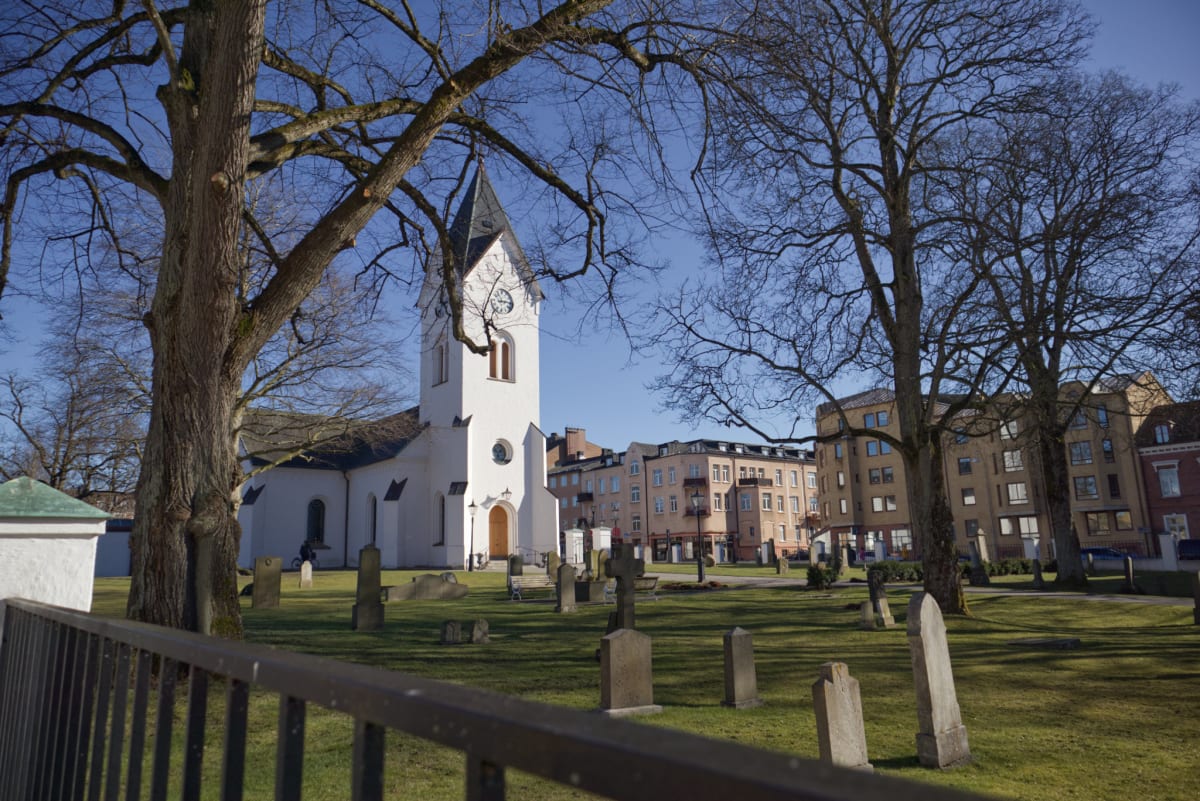 Bilden visar ett landskap med en vit kyrkobyggnad och en grön kyrkogård. Kyrkan har ett högt, spetsigt klocktorn med ett mörkt tak och en synlig urtavla. På kyrkogården, framför kyrkan, finns flera gravstenar utspridda på ett gräsfält. Stora nakna tr