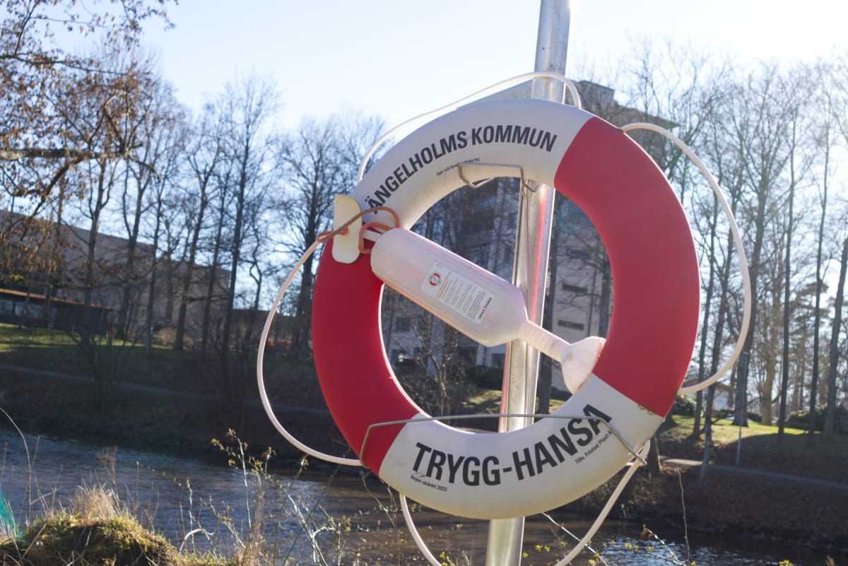 En röd och vit livboj hänger fäst vid en metallstolpe. På livbojen står det skrivet "ÄNGELHOLMS KOMMUN" och "TRYGG-HANSA". En orangefärgad lina är knuten till livbojen, och en liten vit behållare, möjligen en livräddningsfackla eller liten vattentät