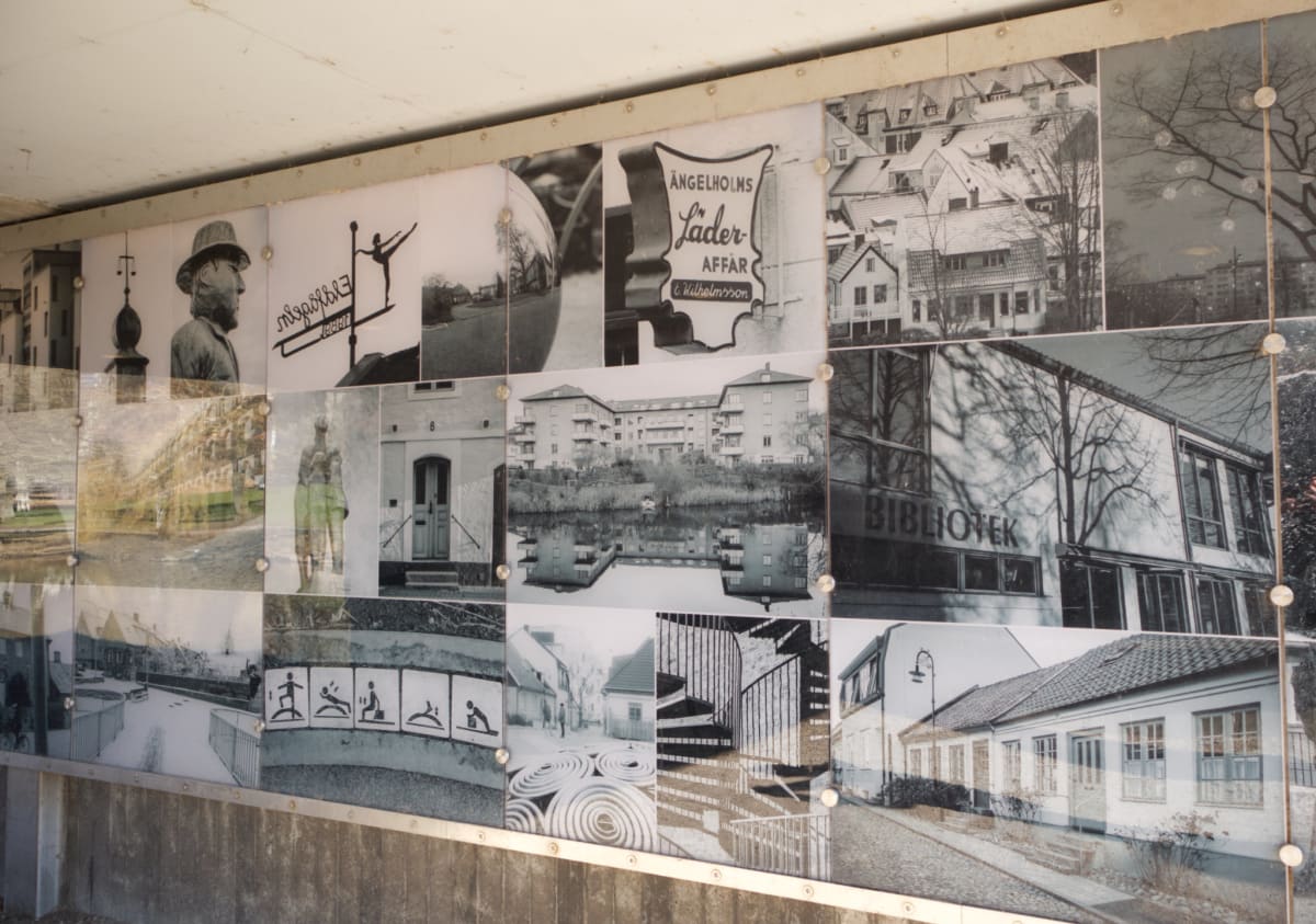 Bilden visar en installation med flera svartvita fotografier monterade på en vertikal yta, möjligtvis en vägg. Fotografierna, som är arrangerade i ett rutmönster, föreställer olika motiv såsom byggnader, gator, ett vattendrag med reflektioner, en sta