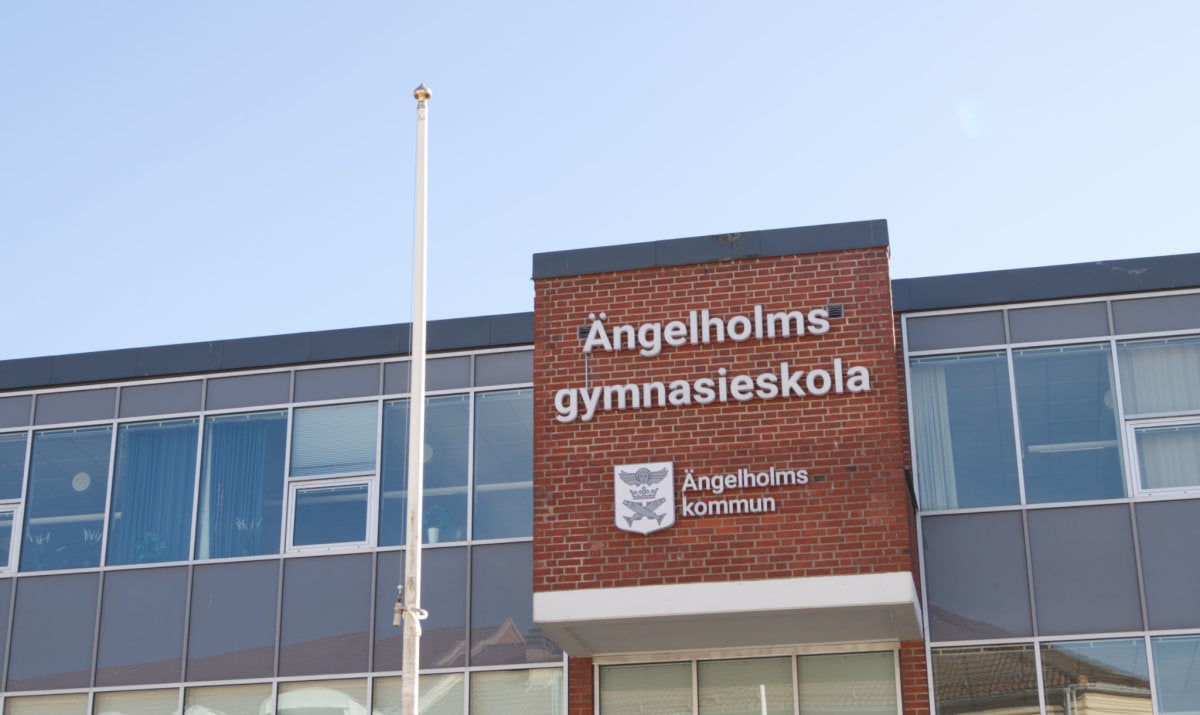 Bilden visar fasaden på en byggnad med texten "Ängelholms gymnasieskola" i vita bokstäver på en tegelvägg. Under texten finns en symbol med två fiskar och en ängel, samt texten "Ängelholms kommun". Till vänster om tegelväggen syns en del av en modern