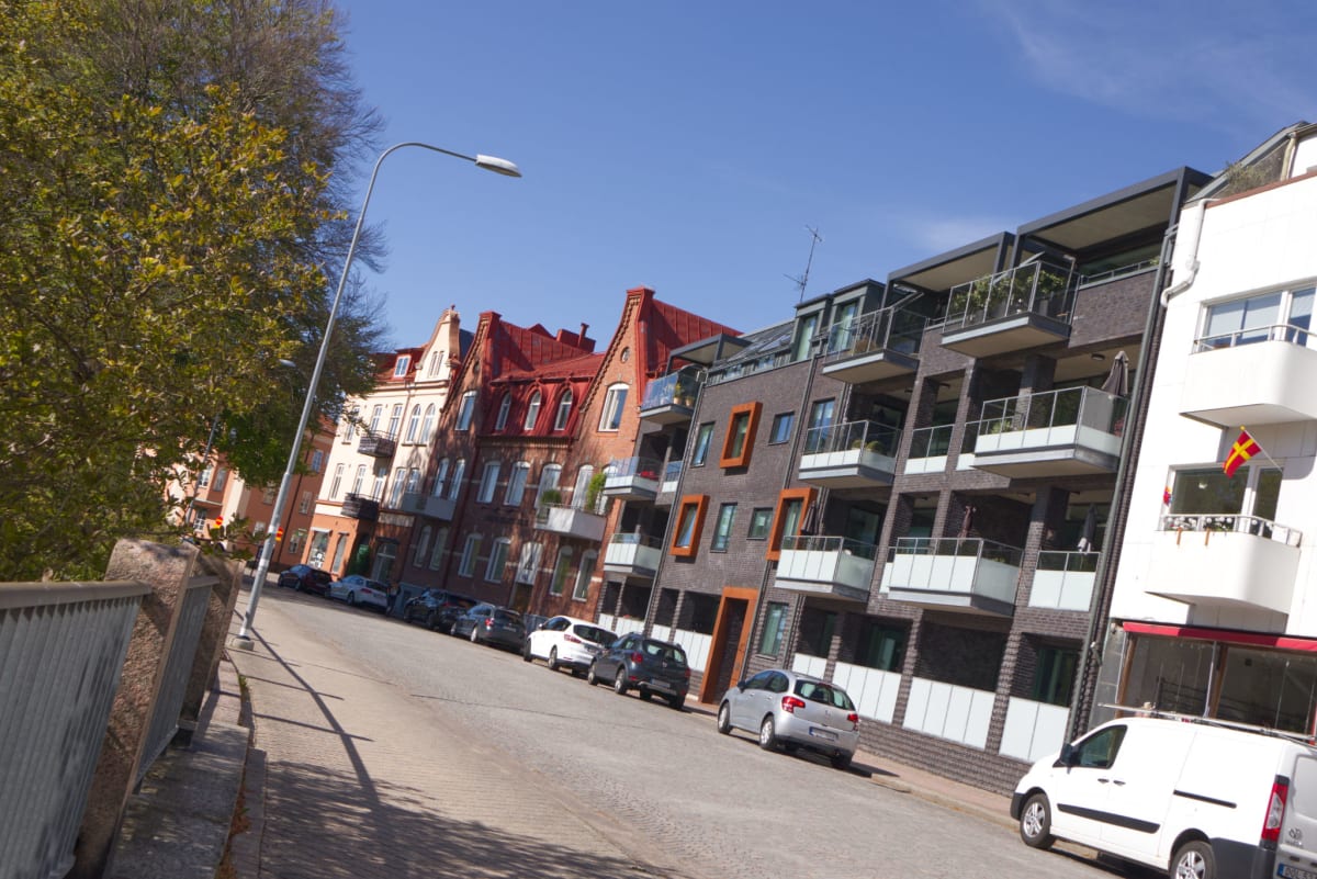 Bilden visar en gata med parkering längs sidan under dagtid. Till vänster syns delar av en mur eller ett räcke samt en grönskande trädtopp som delvis täcker himlen. Längs gatan står flera bilar parkerade. Husen längs gatan varierar i stil; några är ä