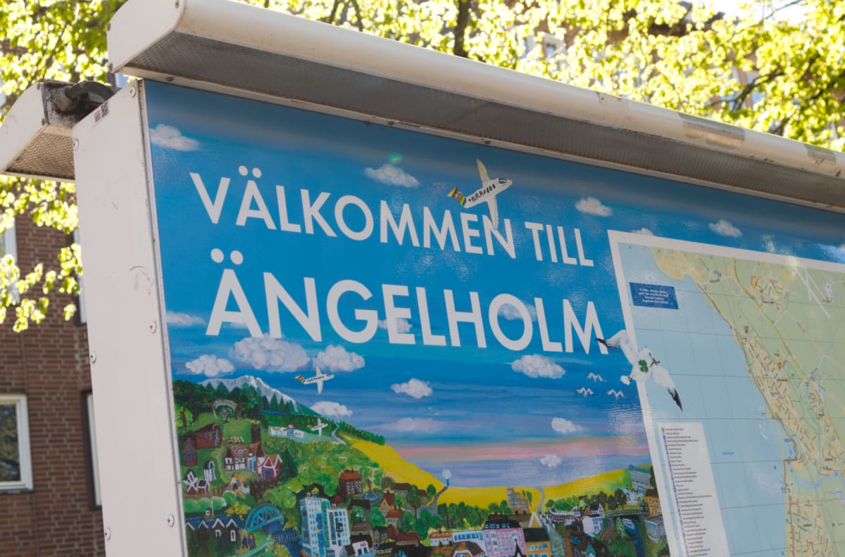 Bilden visar en informationsskylt utomhus med texten "Välkommen till Ängelholm" i vit versalfon på en ljusblå bakgrund. Ovanför texten syns två tecknade flygplan och några moln. Under texten och flygplanen finns en illustrerad landskapsbild med gröna