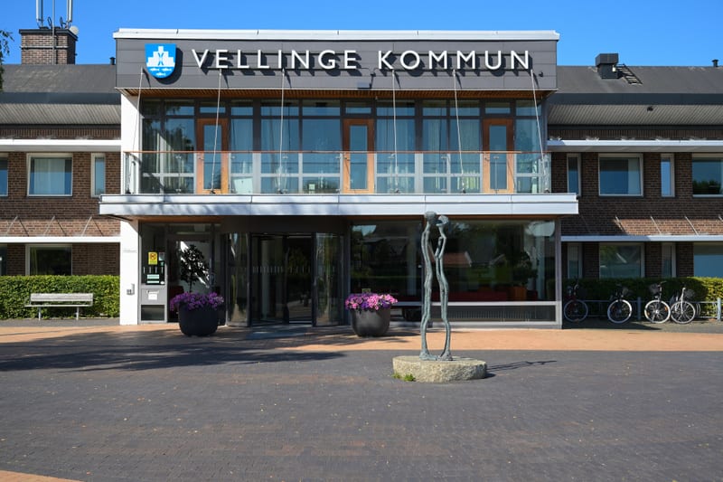 Bilden visar fasaden till Vellinge kommunhus under dagtid. Byggnaden har en modern del med stora glaspartier och en skylt där det står "VELLINGE KOMMUN" med kommunens logotyp. Denna del har också en balkong med glasräcke. På markplan finns en roteran