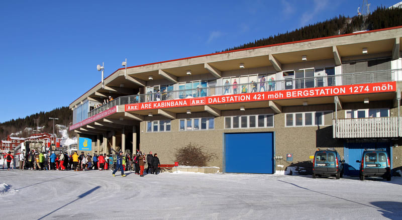 Bilden visar utsidan av en byggnad med en stor röd banderoll som löper längs fasaden. På banderollen står texten "ÅRE KABINBANA DALSTATION 421 möh BERGSTATION 1274 möh". Framför byggnaden står en grupp människor iklädda vinterkläder och med skidor, v