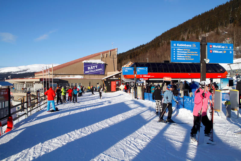 Bilden visar ett snötäckt landskap vid en skidort under dagtid med flera skidåkare och snowboardåkare. I förgrunden syns skuggor som sträcker sig över snön. Till vänster finns en byggnad med texten "RÄTT INSTÄLLNING" på en blå skylt. Flera personer ä