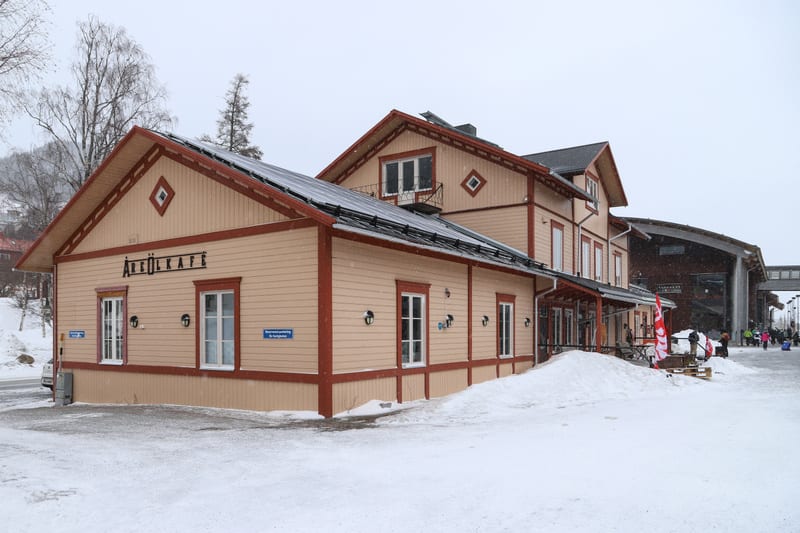 En beige byggnad med röda lister och fönsterkarmar står täckt av snö. På byggnadens tak finns solpaneler. På fasaden syns texten "ÅREÖL KAFE" ovanför två vita fönster. Under fönstren sitter utomhuslampor. Framför byggnaden ligger snöhögar och det snö