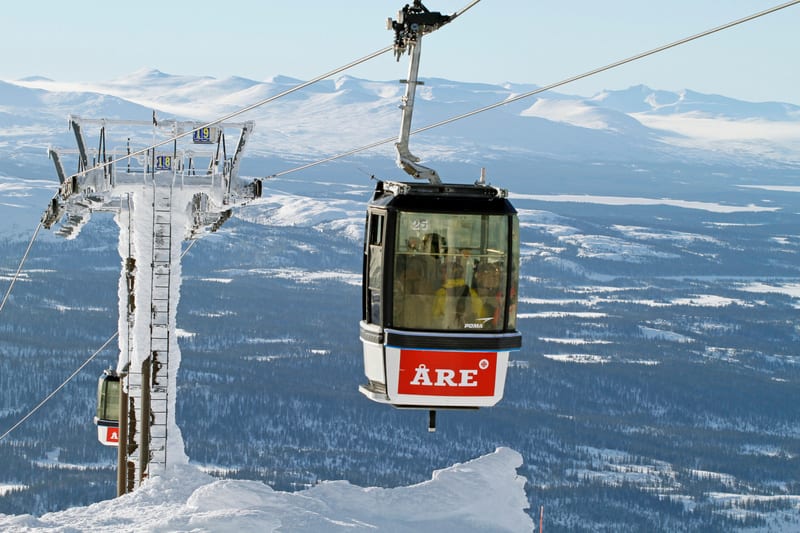 Bilden visar en gondollift med en enskild gondol i förgrunden, som är transparent med ett rött fält undertill med texten 'ÅRE'. Gondolen är i rörelse och ser ut att ha passagerare inuti. Bredvid gondolen, till vänster i bilden, finns en isbeklädd lif