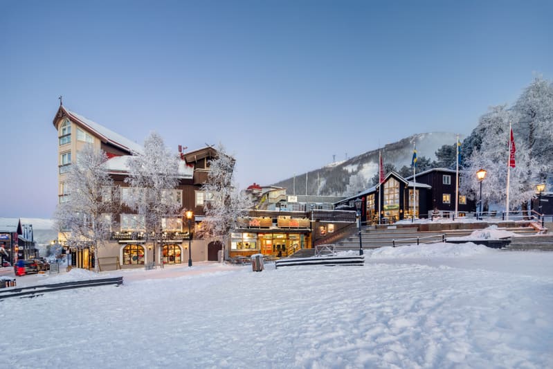 En vinterbild från en skidort som visar en bebyggd dalgång med flertalet byggnader, träd täckta av snö och frost, samt delvis snötäckta berg i bakgrunden. Ett stort snötäckt öppet område finns i förgrunden. Till vänster i bilden syns en stor byggnad