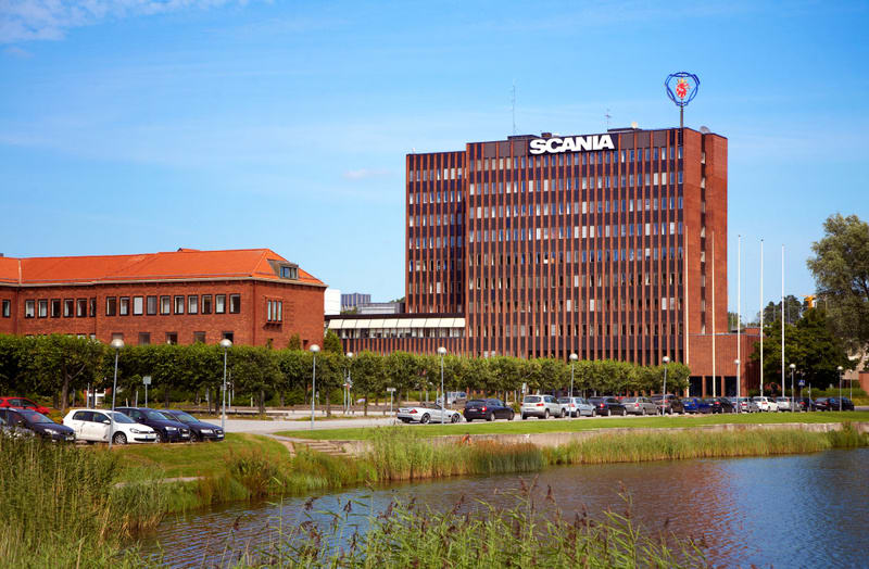 Bilden visar fasaden på en hög, rektangulär byggnad med rödbrunt tegel och många vertikala fönsterrader. Ovanpå byggnaden syns texten "SCANIA" i stora, vita bokstäver samt en emblem som liknar Scanias logotyp. Framför denna byggnad finns en lägre teg