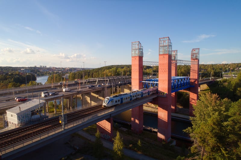 Bilden visar ett landskap med flera broar under klarblå himmel. I förgrunden syns en lång bro med järnvägsspår där ett blått och vitt tåg är på väg att passera. Bredvid tågbron finns fyra kraftiga, rödfärgade pelare med glasstrukturer på toppen, samm