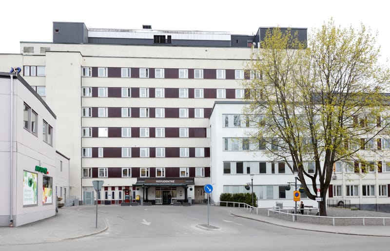 Bilden visar utsidan av ett stort byggnadskomplex, möjligen ett sjukhus eller en vårdcentral, med skylten 'HUVUDENTRÉ' ovanför en av ingångarna. Byggnaden har flera våningar med många fönster och är huvudsakligen vit eller ljusbeige med bruna detalje