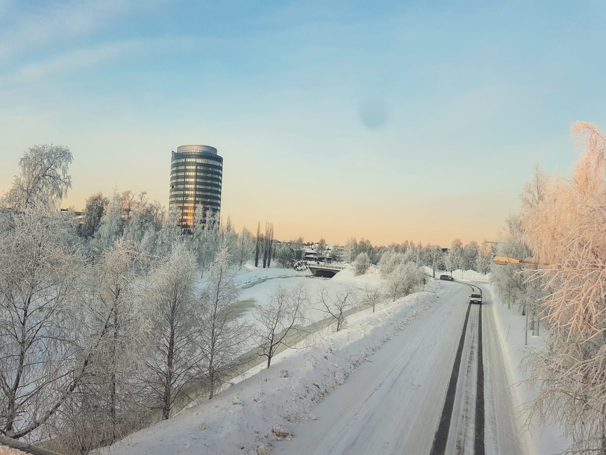Bilden visar ett vinterlandskap med en väg i förgrunden, täckt av snö och med tydliga spår från fordon. Till vänster om vägen finns en snötäckt mark och en delvis frusen vattensamling eller å, omgiven av träd och buskar som är täckta av frost eller s