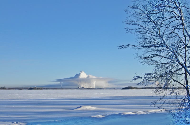 Bilden visar ett snötäckt, fruset landskap under en klarblå himmel. I förgrunden syns en stor yta täckt av snö, som kan vara en frusen sjö eller ett öppet fält, med långa skuggor som sträcker sig över ytan. Till höger i bilden syns grenar från ett lö