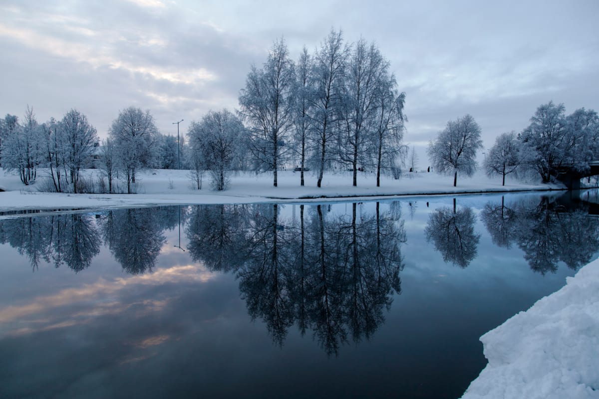 En vinterlandskapsbild som visar en vattensamling, troligen en å eller flod, med snöklädda stränder och träd. Vattnet reflekterar den molniga himlen och de snötäckta träden längs strandkanten. Träden är täckta av vit frost eller snö. På höger sida i