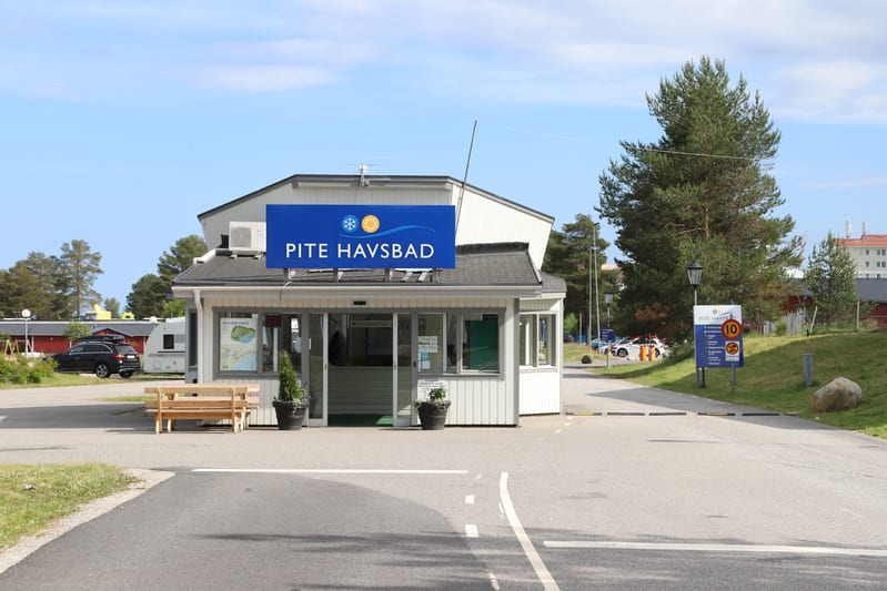 Bilden visar entréområdet till "Pite Havsbad" under dagtid, med en reception eller servicebyggnad i centrum. Byggnaden är vit med grått tak och har ett stort blått skylt med texten "PITE HAVSBAD" och två symboler. Framför byggnaden står en träbänk oc