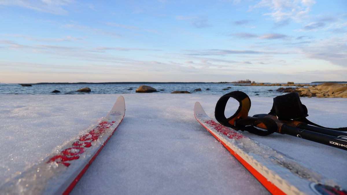 Bilden visar ett par längdskidor och stavar som ligger på ett istäckt, snöigt underlag vid en vattenkant. I bakgrunden syns ett lugnt vattenområde, möjligen ett hav eller en stor sjö, med flera stenar eller små öar utspridda. Vattnet verkar vara frit