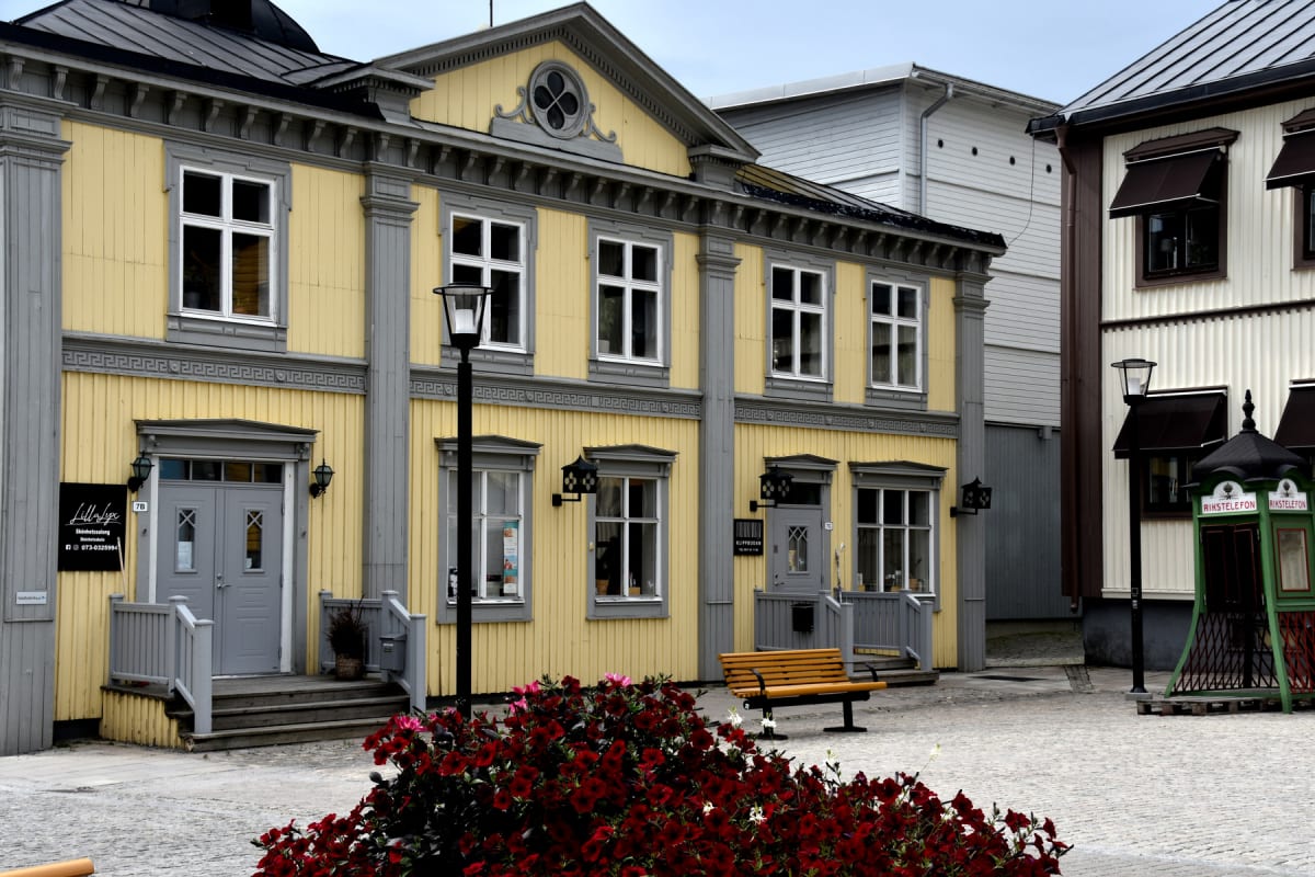 En bild som visar en kullerstensbelagd gata, med två byggnader i fokus. Till vänster finns en gul träbyggnad med vita fönster och grå detaljer. Träbyggnaden har flera fönster i två våningar, en altan med trappor och entrédörrar som är grå. På altanen