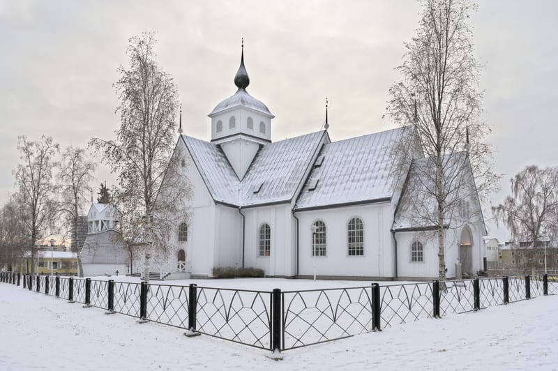 Bilden visar en vit kyrkobyggnad sedd från en sidovinkel under vintertid. Kyrkan är huvudsakligen vitmålad med mörka detaljer på taket och en svart spira med en kula överst. Taket är täckt av ett tunt lager snö. Runt kyrkan syns snötäckta markytor oc
