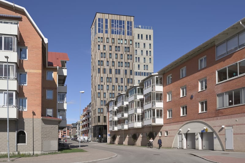 En gata inramad av byggnader under en klarblå himmel. På vänster sida syns ett tegelfärgat hus med vita balkonger och fönster. Delar av huset har en grå stengrund och en röd takdel syns ovanför ett intilliggande byggnadsparti. I mitten av bilden domi