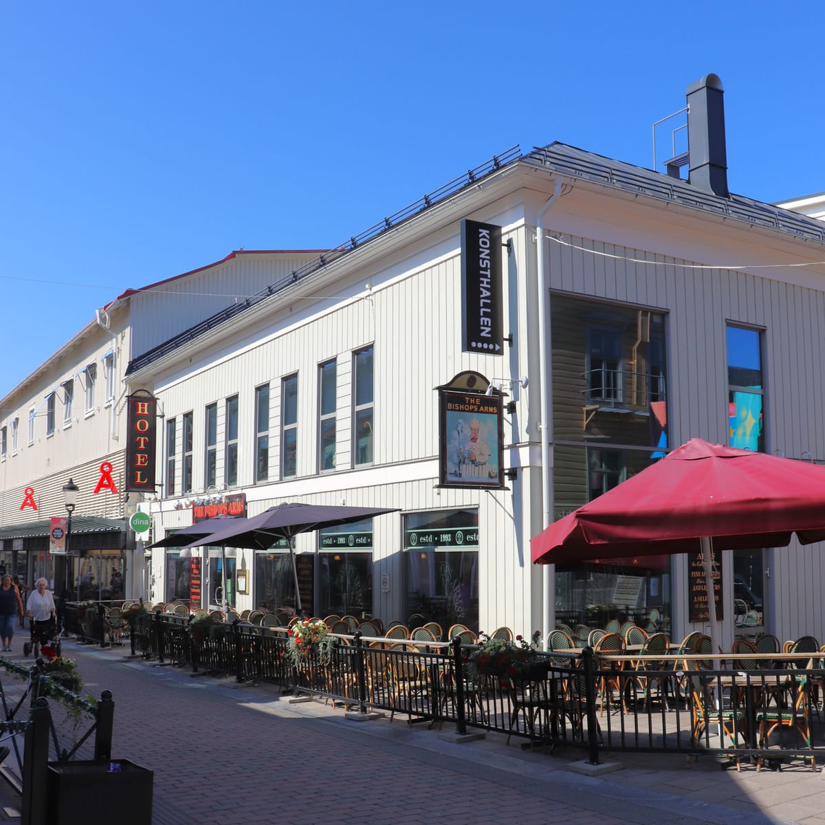 Bilden visar fasaden på en vit byggnad med flera skyltar. En skylt, vertikalt placerad, lyder "KONSTHALLEN" med tre punkter under texten. En annan skylt är "THE BISHOPS ARMS" med en bild under texten. Byggnaden har många fönster och en svart skorsten