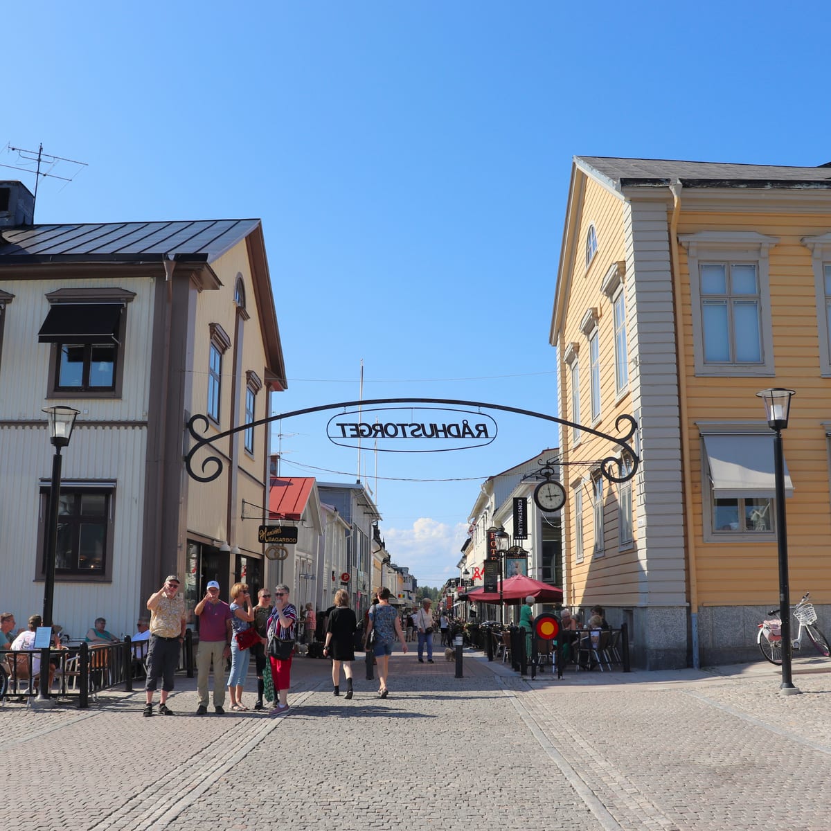 Bilden visar en gata i en stad på en solig dag. Gatorna är kullerstensbelagda och kantas av pastellfärgade byggnader i traditionell stil, främst gult och beige. Flera personer, både grupper och enskilda individer, går på gatan eller sitter vid uteser