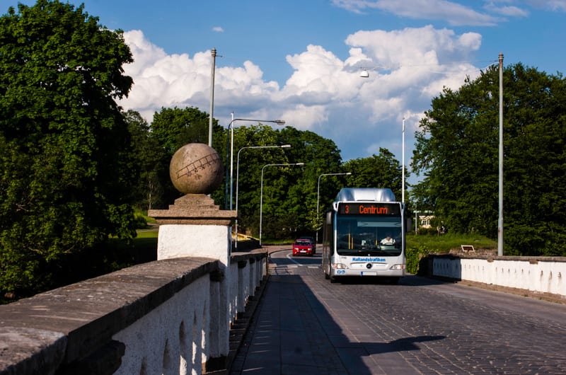 Bilden visar en buss och en personbil på en väg som kantas av grönska och ett betongräcke. Bussen är vit med texten "Hallandstrafiken" och destinationsskylten "3 Centrum". En man kan ses bakom ratten på bussen. Bredvid bussen syns en röd personbil. V
