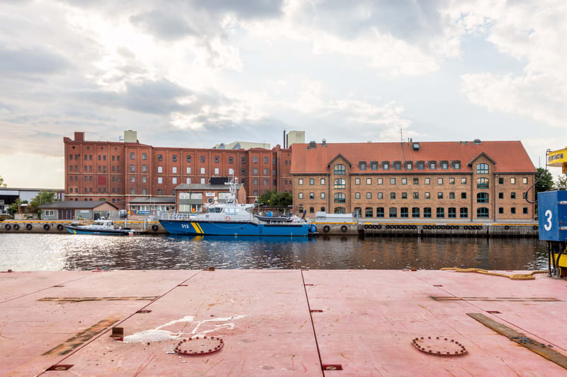 Bilden visar ett vattendrag med flera byggnader i bakgrunden. I förgrunden syns en stor, rödaktig yta, förmodligen ett kajområde eller däck på ett fartyg. Vid kajen ligger två båtar. Den ena är en större blå och vit båt med texten "312" och en gul ra
