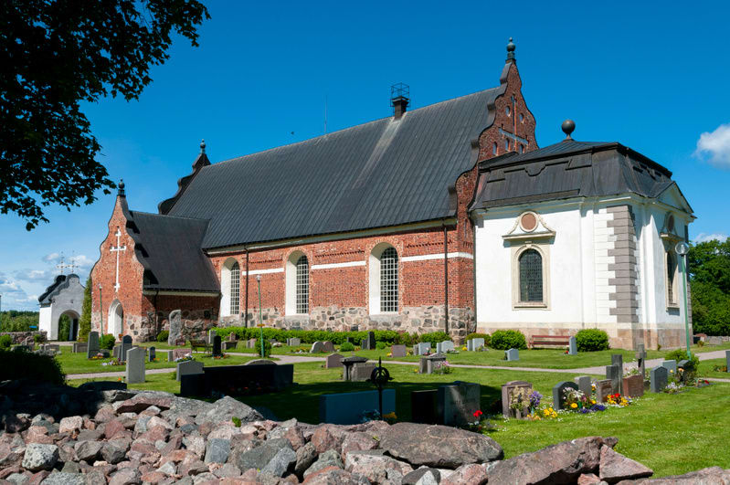 En bild som visar en kyrka med en kyrkogård i förgrunden och på sidorna. Kyrkan är byggd av rött tegel med ett mörkt tak, och har en vit byggnadskropp fäst vid sig. En del av kyrkan har en vit korssymbol på sin gavel. Längst ner i bilden syns en oreg
