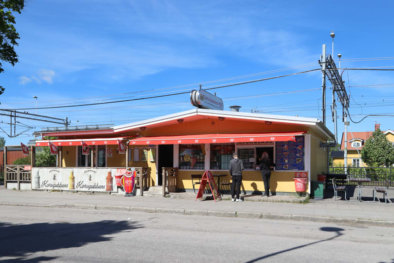 Bilden visar en gul kioskbyggnad, troligen en korv och glasskiosk, med en röd markis som sträcker sig längs framsidan. Flera röda flaggor med texten "Originalet Korvgubbens" hänger under markisen. På taket finns en skylt med texten "KORVGUBBENS" och